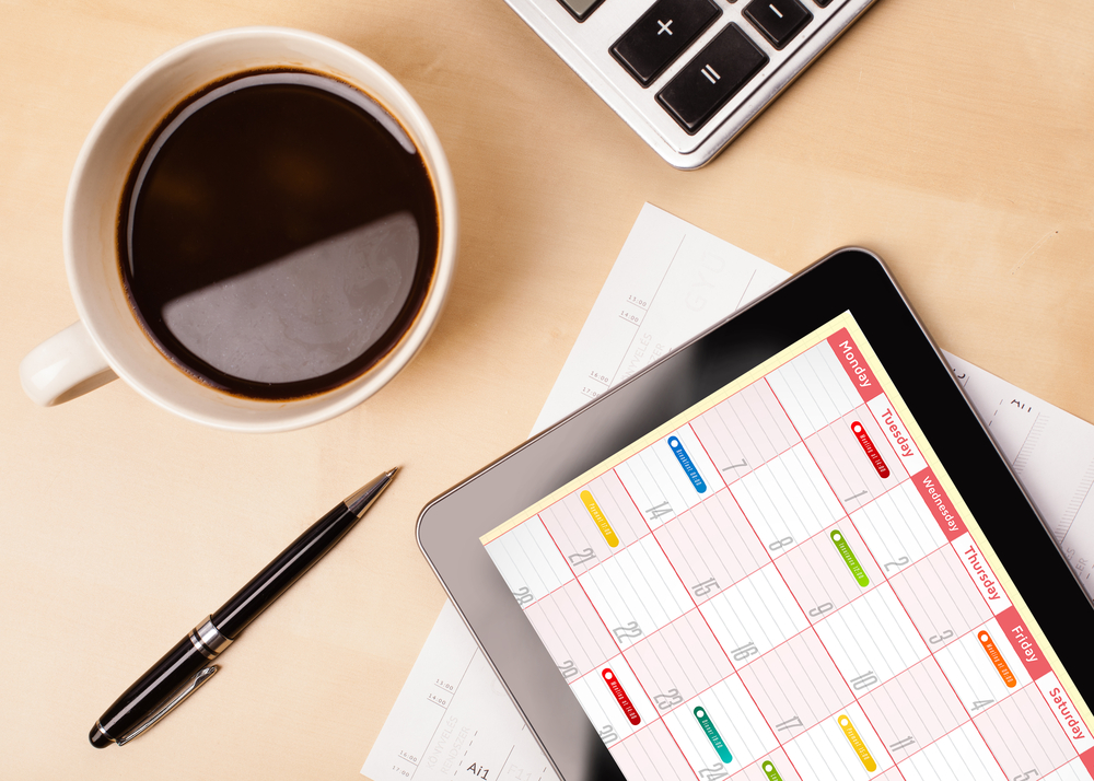 Workplace with tablet showing calendar and a cup of coffee on a wooden work table